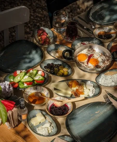 Güneş ışığında, küçük tabaklarla kurulmuş serpme kahvaltı sofrası: yumurta, zeytin, peynir ve taze sebzeler